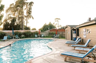 Outdoor pool area with lounge chairs, trees, and a Pepsi machine on a sunny day.
