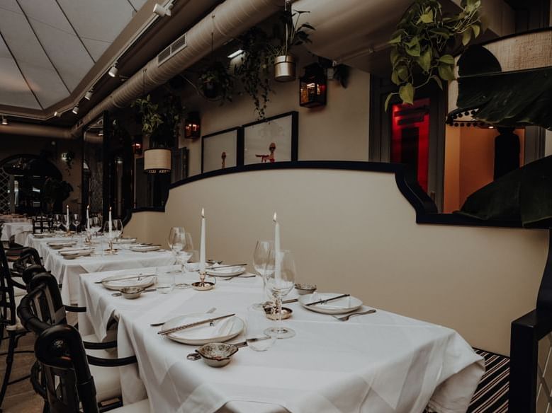 An elegant dining area with white tablecloths, lit candles, and modern chairs set against a wall with framed art and hanging plants.