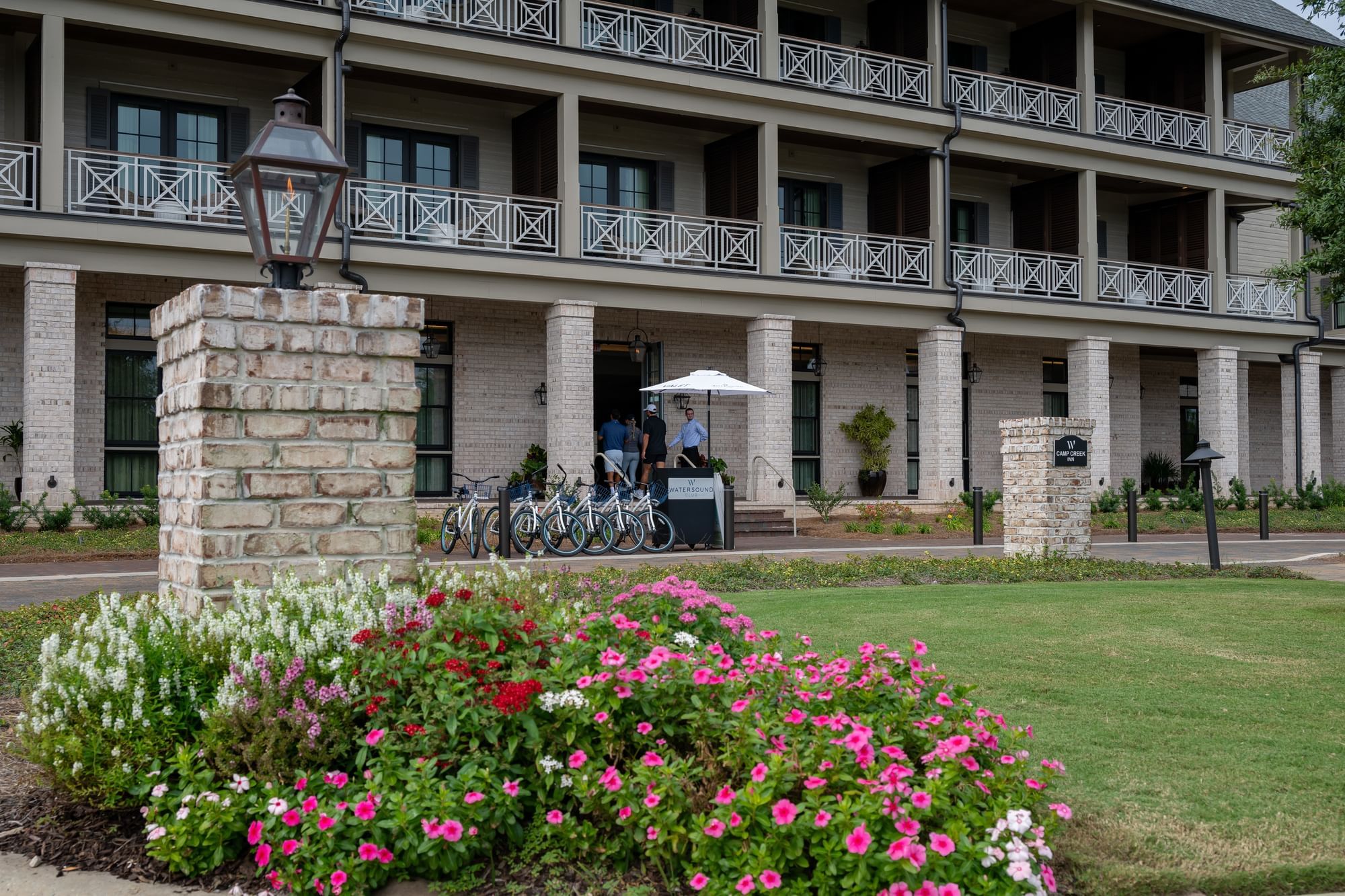 Large building with many balconies, bicycles, and people under an umbrella, and a colorful flower bed in front.