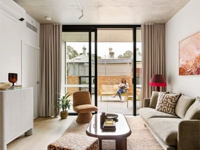 Woman reading on a balcony through a sliding glass door in a cozy apartment.
