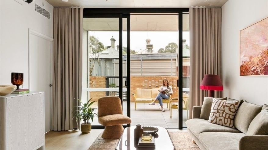 Woman reading on a balcony through a sliding glass door in a cozy apartment.