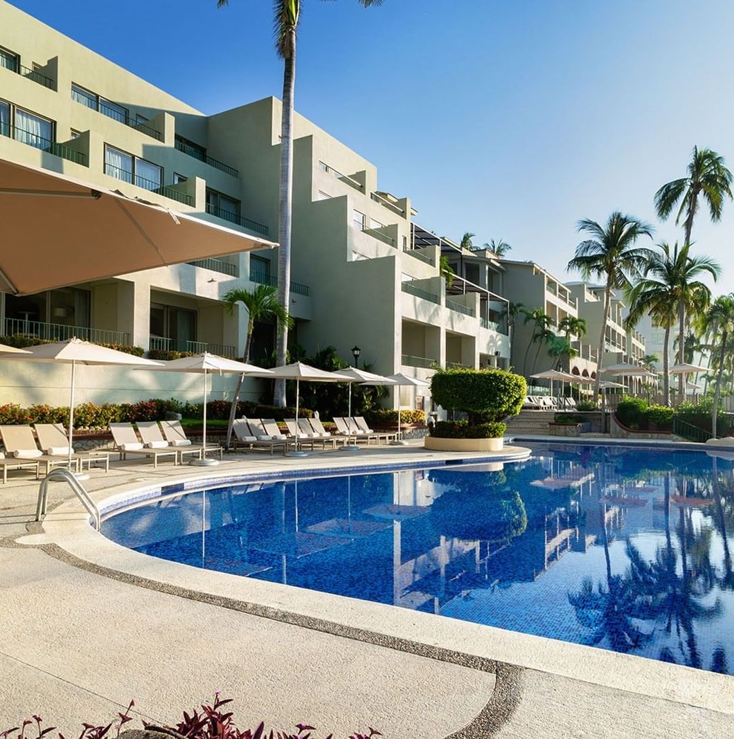 Luxury swimming pool with lounge chairs and palm trees under a blue sky at Camino Real Acapulco Diamante