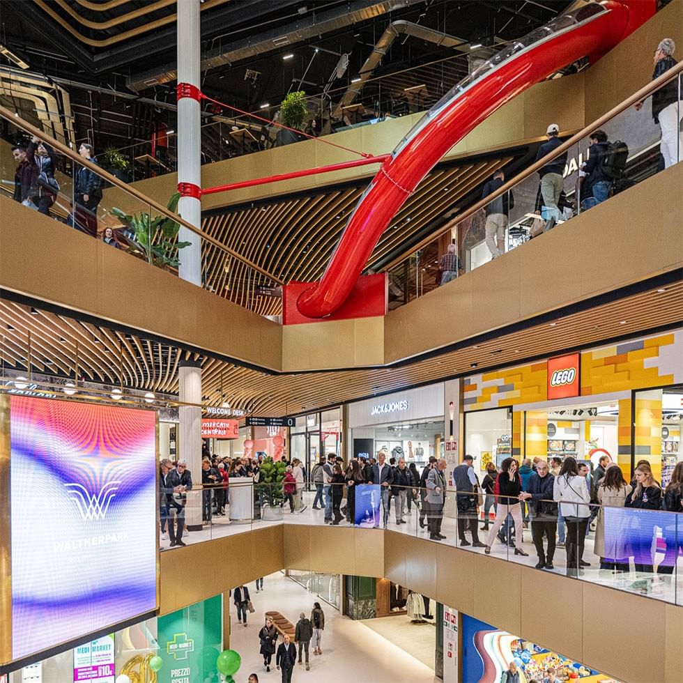 Crowded mall with red curved structure, people on balconies, and large screens displaying advertisements.