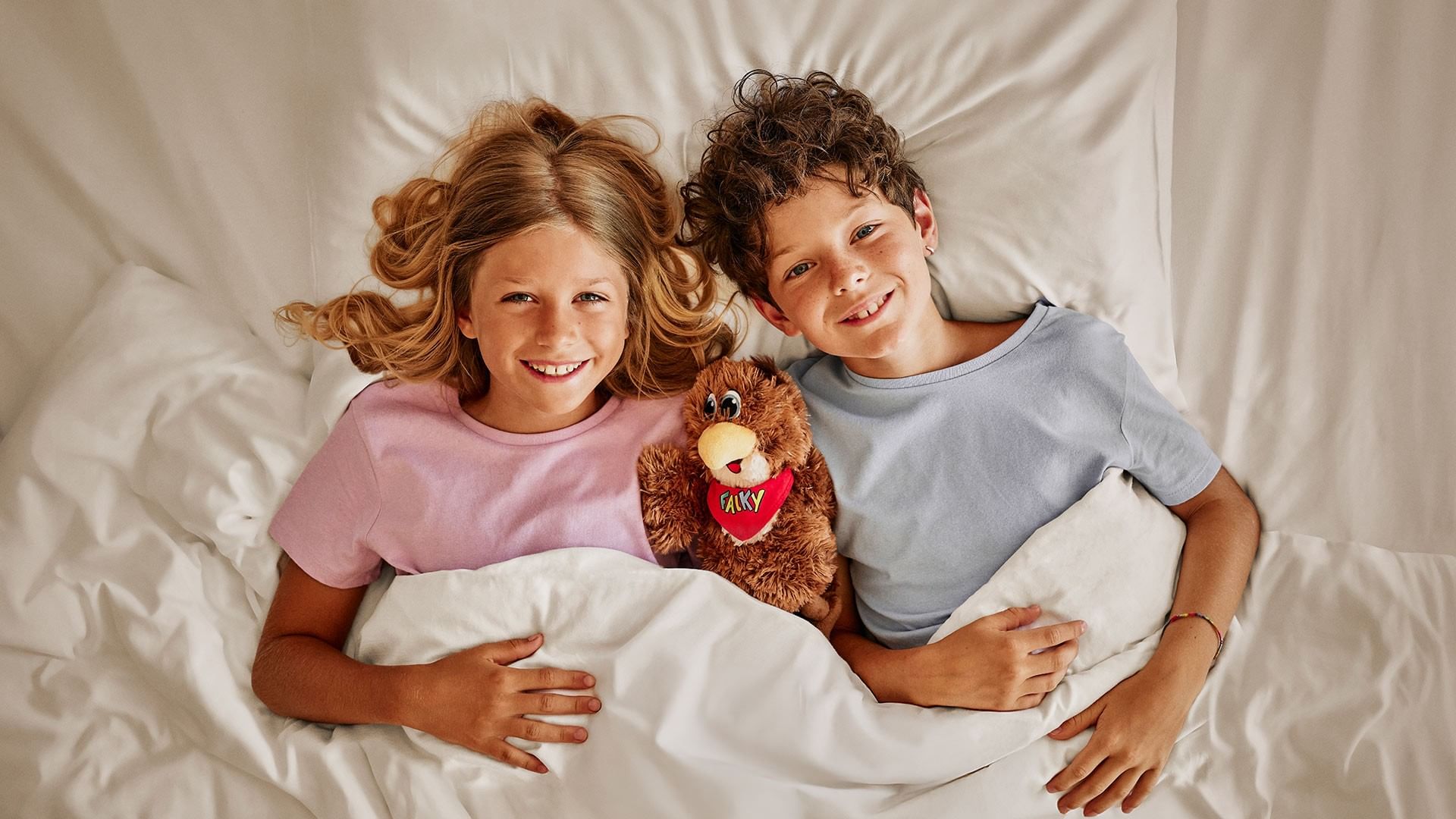 Geschwister liegen im Bett, halten eine Puppe und lächeln in die Kamera.