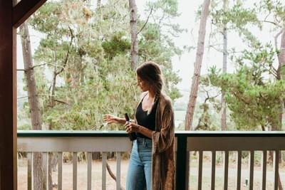 Woman on porch holding wine glass and bottle, surrounded by tall trees.
