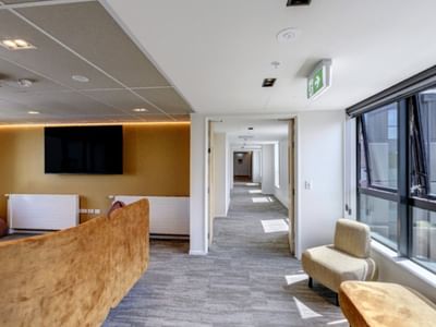 Modern hotel lounge area with wooden reception desk, wall-mounted TV, seating, and bright corridor view in Tupuārangi.