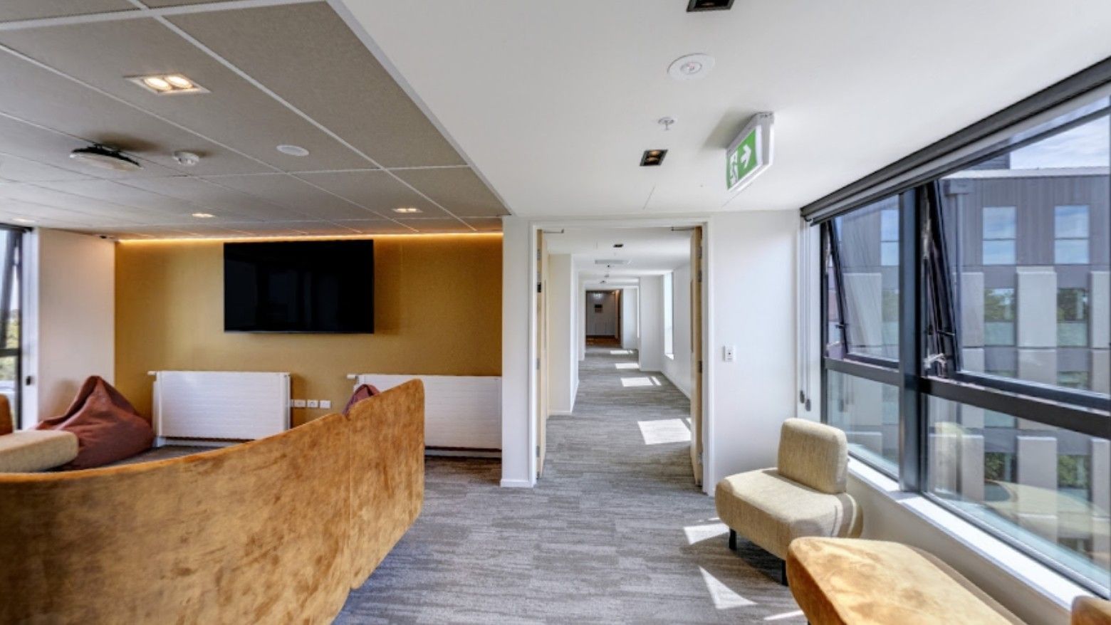 Modern hotel lounge area with wooden reception desk, wall-mounted TV, seating, and bright corridor view in Tupuārangi.