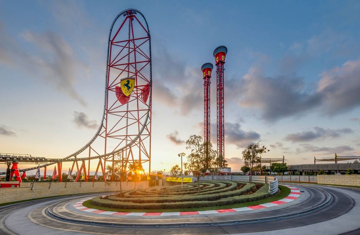 Red Ferrari Land roller coaster and drop towers at sunset, with a curved racetrack in the foreground at PortAventura World.