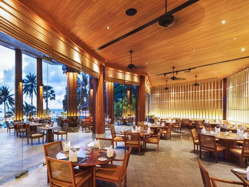 Beach Restaurant at The Surin Phuket, with wooden decor and palm trees visible through large glass windows