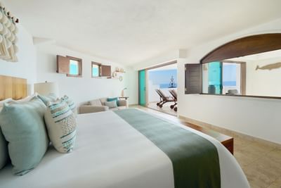 A bedroom with a large bed, pillows, and a couch facing an open balcony with ocean view.