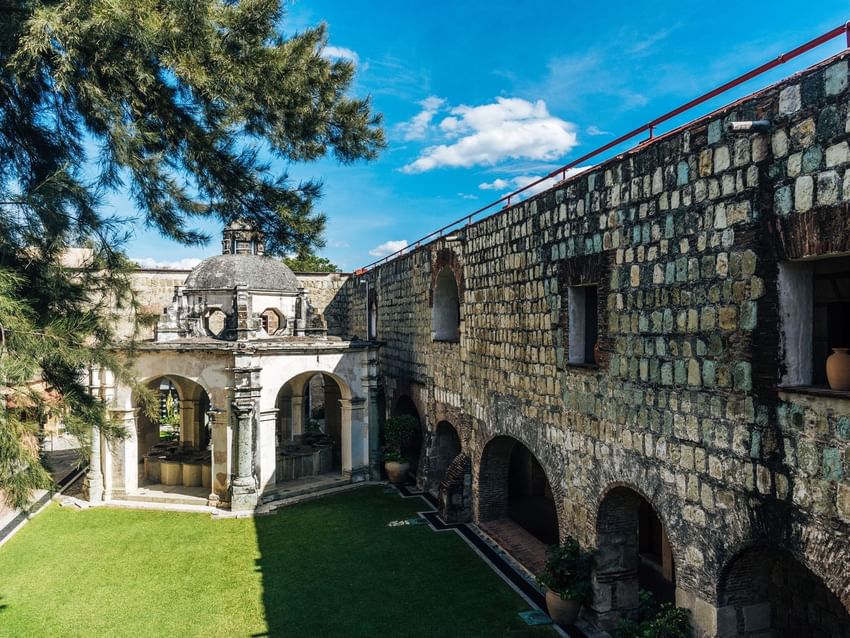 Stone courtyard with an arched pavilion, lush green lawn, and a large tree at Jardín Los Lavaderos in Quinta Real Oaxaca