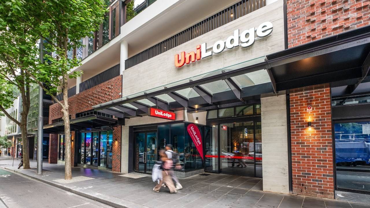 Two people walking past a UniLodge building with brick and glass facade and red banner.