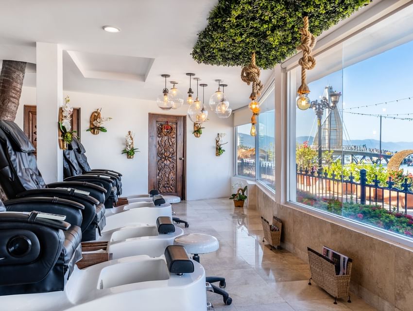 Black pedicure spa chairs facing a large window with ocean pier view and decorative lights inside Los Arcos Suites