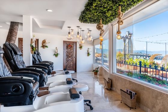Black pedicure spa chairs facing a large window with ocean pier view and decorative lights inside Los Arcos Suites