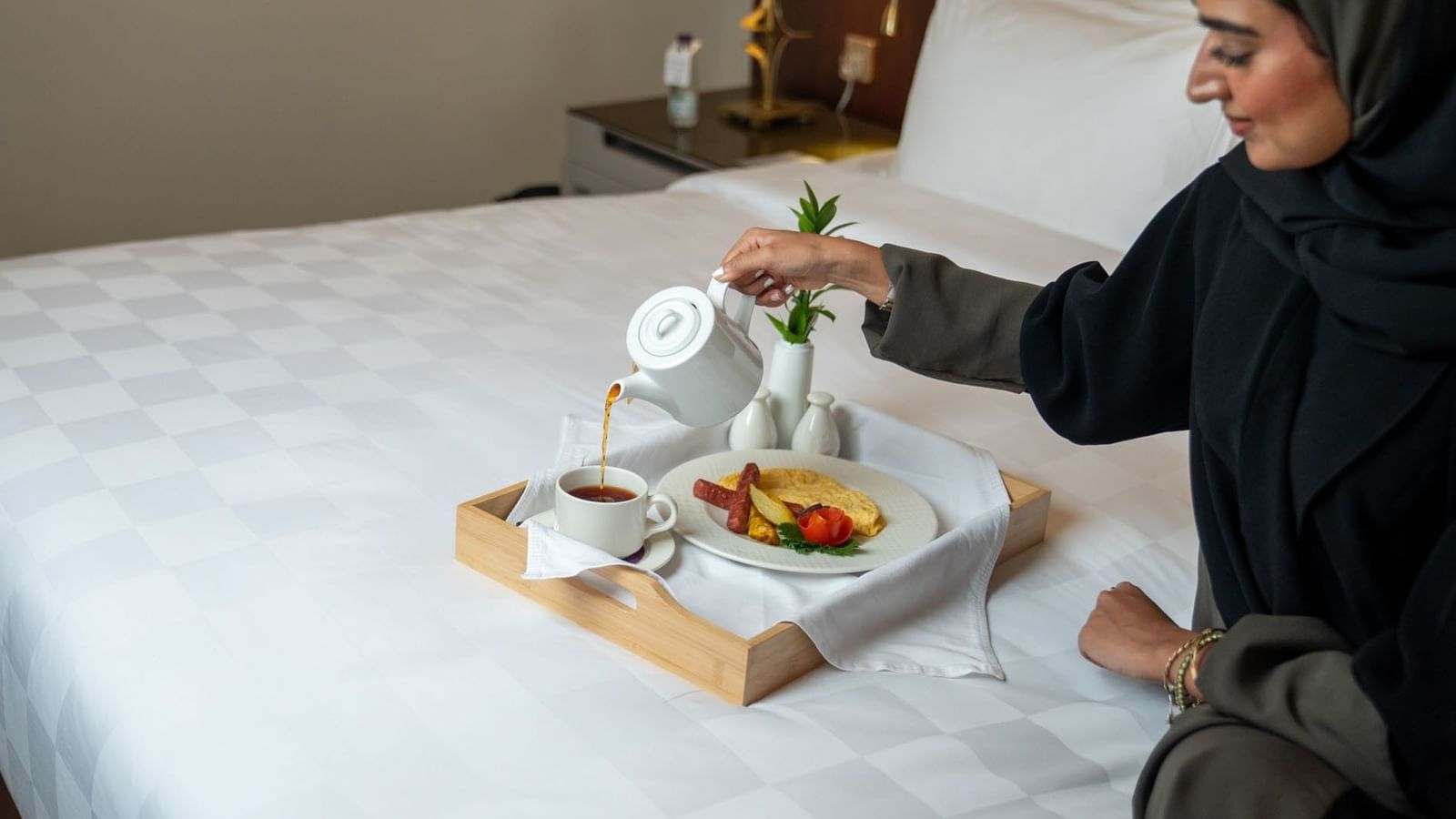 Woman in an abaya pouring tea from a white teapot during breakfast in a hotel room at Saja Warwick Madinah