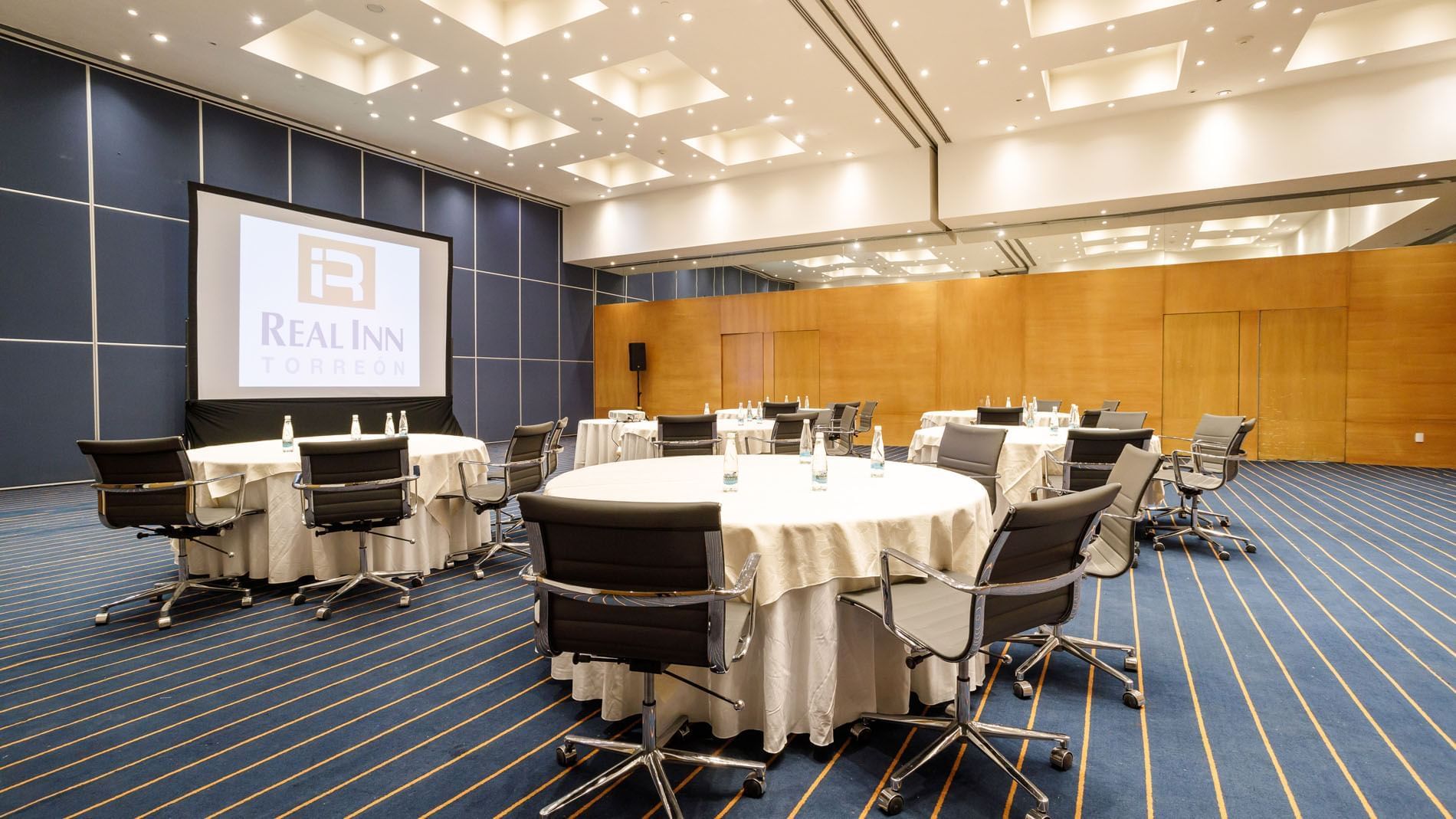 Buenos Aires meeting room with round tables, black chairs, and a large logo projection screen at Real Inn Torreon