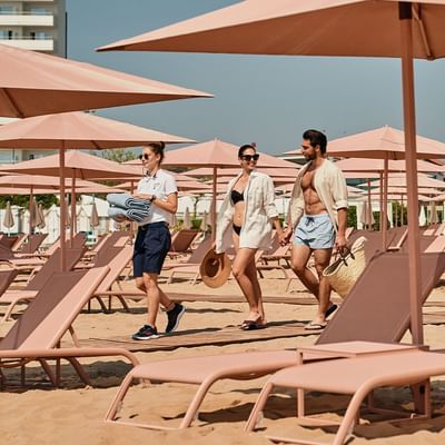 Couple walks hand-in-hand on a sandy beach near Falkensteiner Hotel & Spa Jesolo, passing pink umbrellas and sun loungers