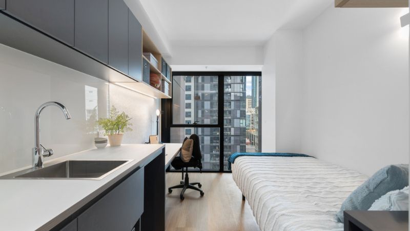 Studio apartment with a bed, desk, chair, and kitchen sink with a city view.