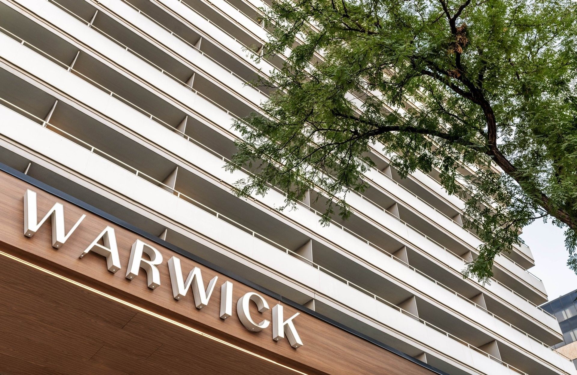 White hotel balconies by a leafy tree under a clear sky near a wood sign near Warwick Denver