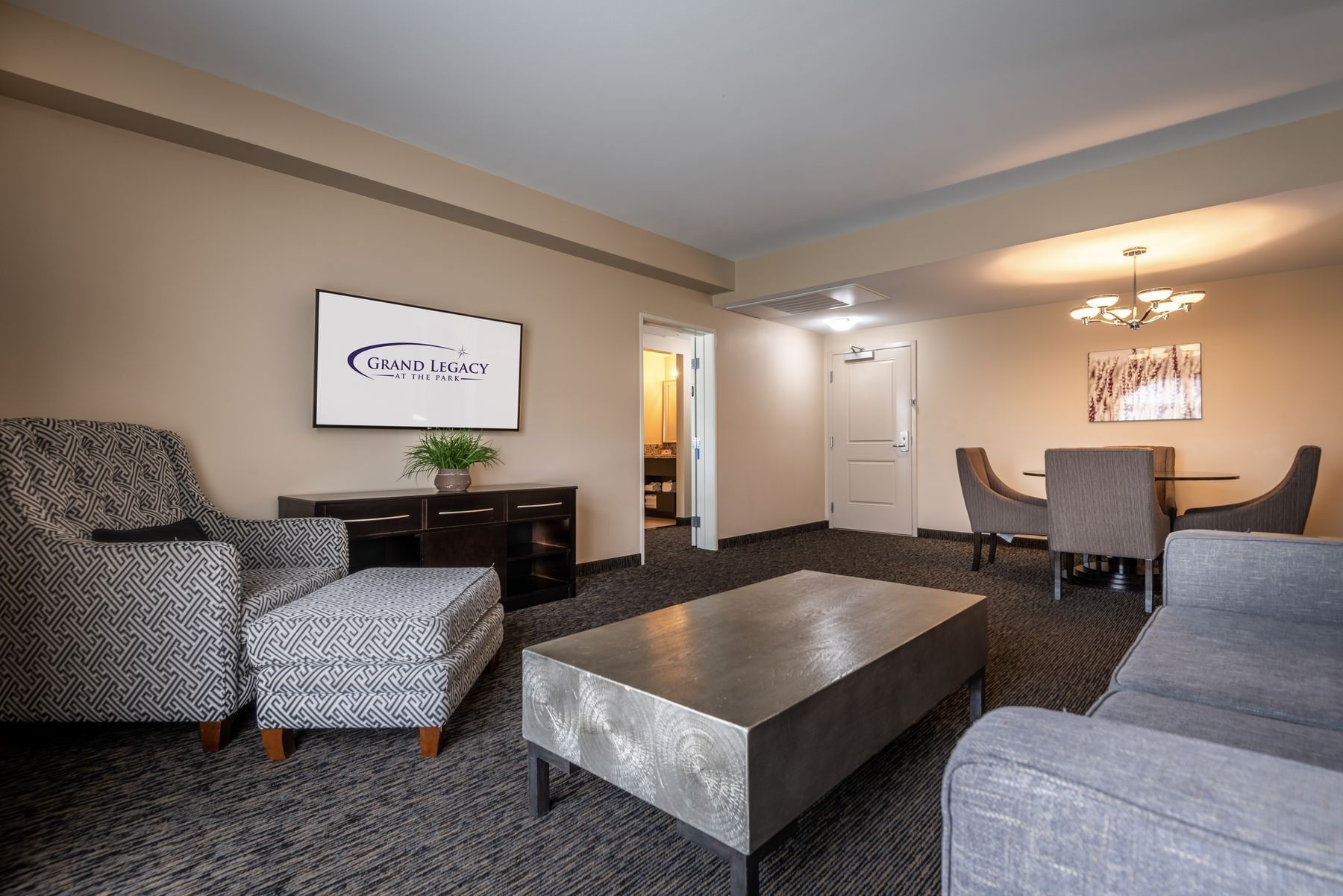 A wide view of a hotel suite's living area with a TV, sofa, armchair, coffee table, and dining table.