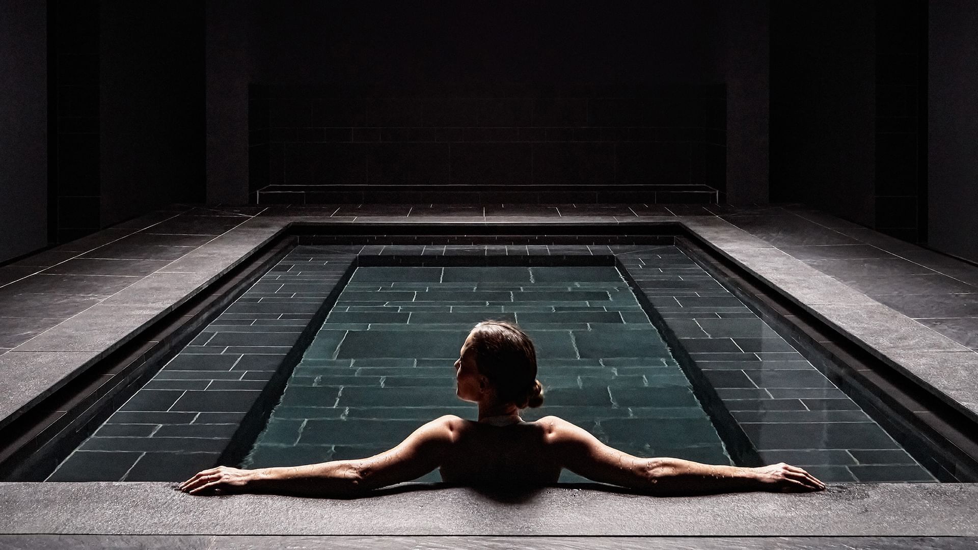 Woman relaxing in a dimly-lit modern spa pool surrounded by dark stone tiles at Falkensteiner Hotel & Spa Iadera