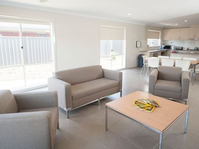 Three gray armchairs, a wooden coffee table, and an open kitchen area in La Trobe University Regional Housing – Shepparton.