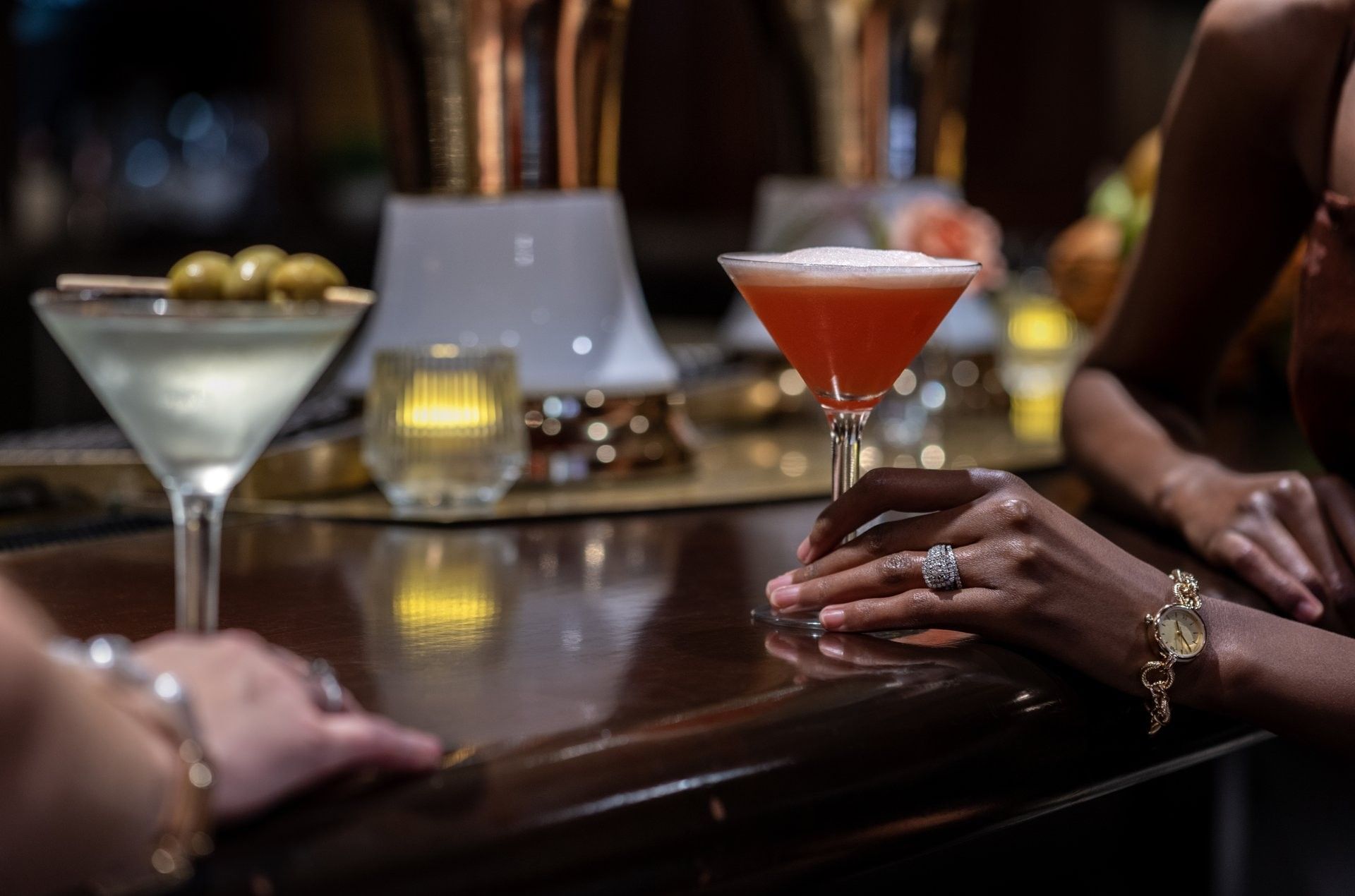 Guest holding a red cocktail by a martini glass on a dark wooden bar counter in Margaux Lounge Bar at Warwick Seattle