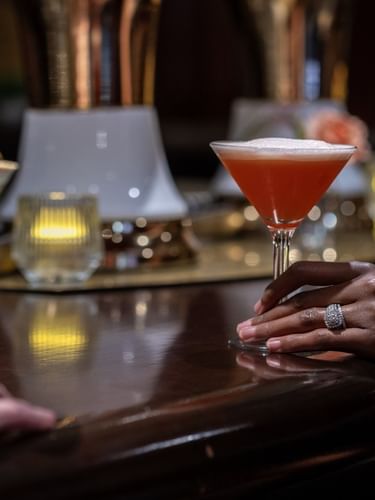 Guest holding a red cocktail by a martini glass on a dark wooden bar counter in Margaux Lounge Bar at Warwick Seattle
