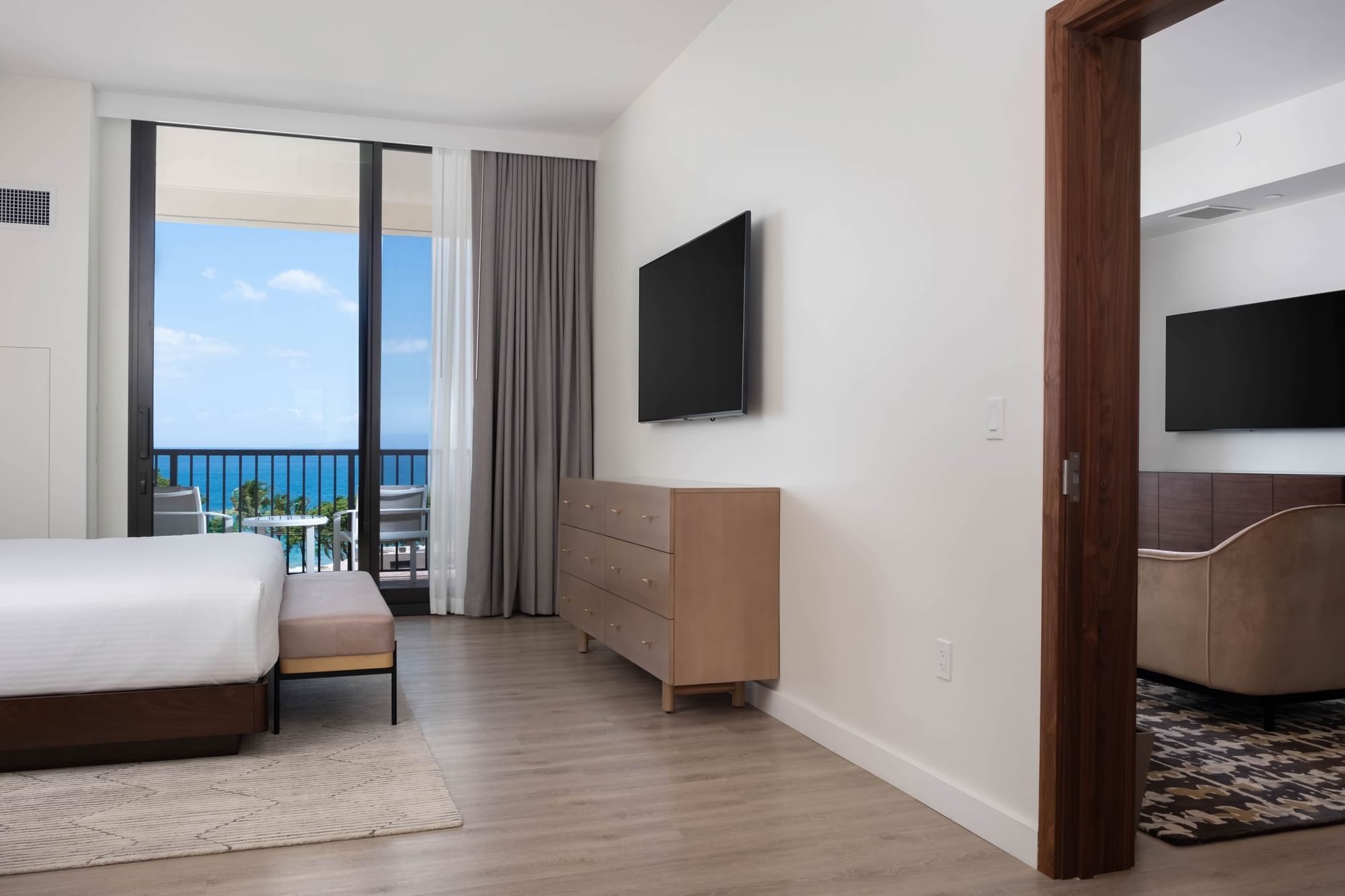 A wide view of a hotel room with a king-sized bed, dresser, flat-screen TV, and balcony with ocean view.