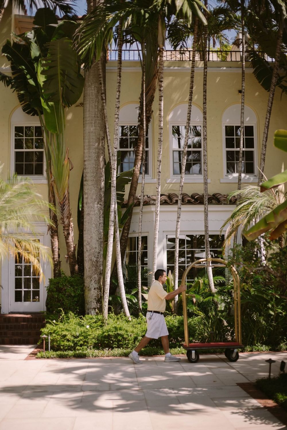 Hotel bellhop pushing brass luggage cart past tall palm trees near courtyard building exterior at The Brazilian Court