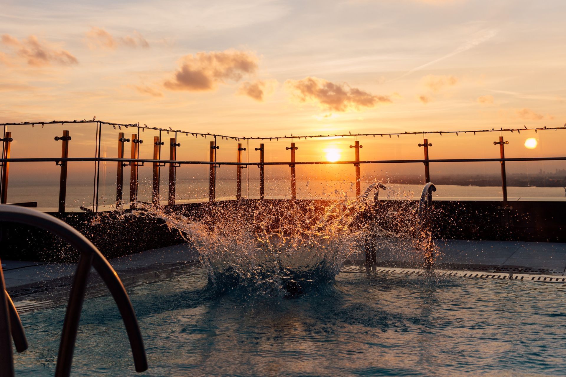 Water splashing high inside a rooftop swimming pool against a glass railing at sunset facing Hotel X Toronto
