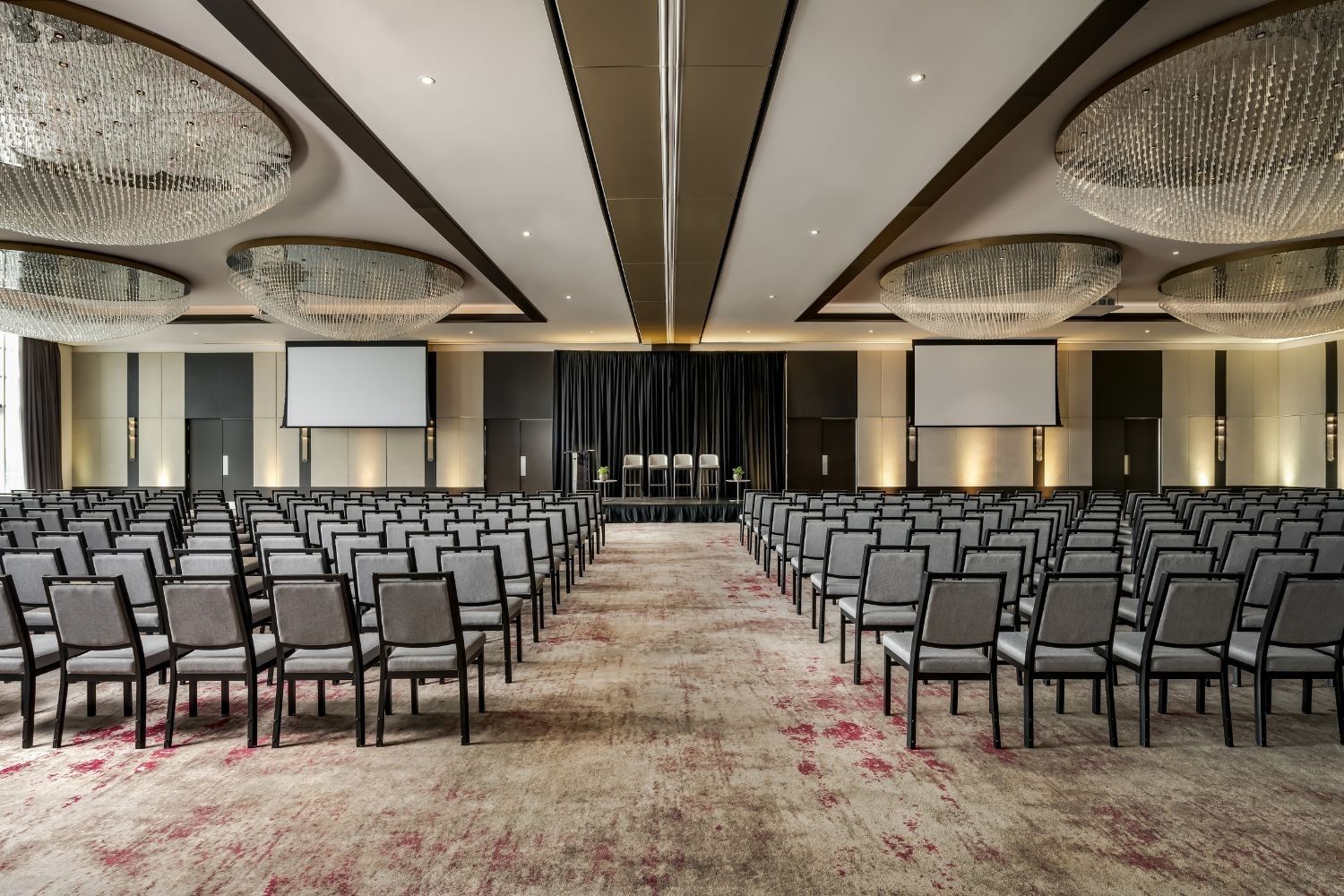 Victory Ballroom with rows of chairs facing a stage and two projection screens beneath chandeliers at Hotel X Toronto