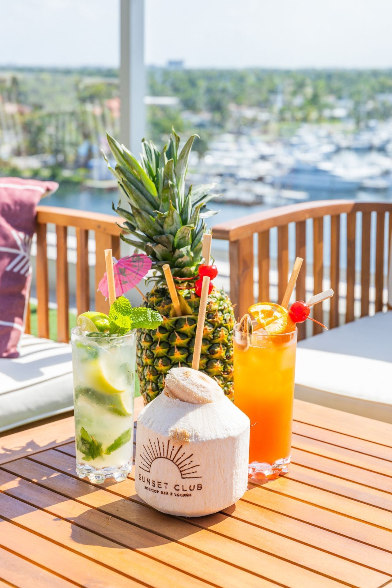 Three cocktails and a coconut on a wooden table with a marina view.