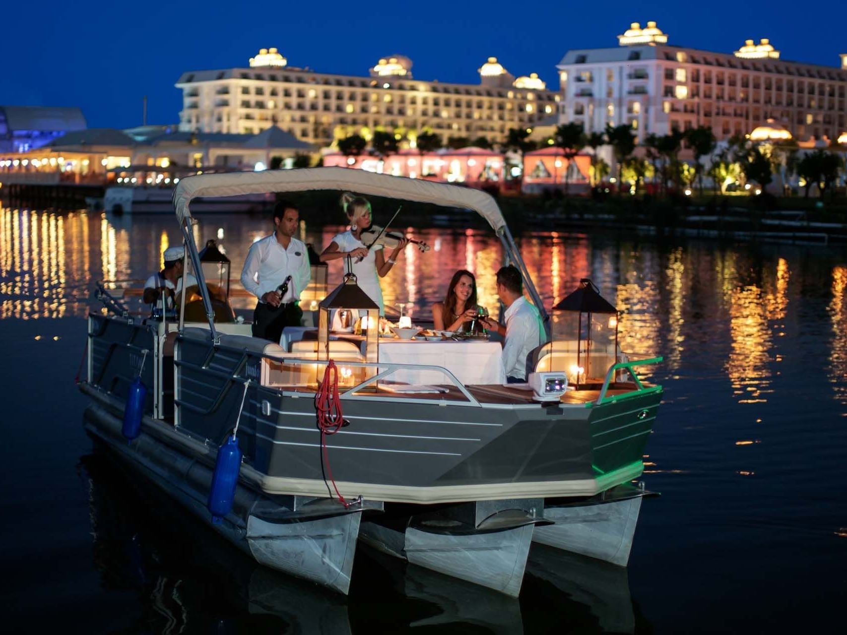 Dinner cruise boat with a couple and a waiter on a calm river with lit-up Titanic Deluxe Golf Belek in the background