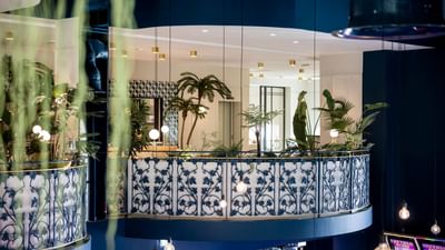 A balcony with blue railings, tropical plants, and ceiling lights, overlooking a gaming area.