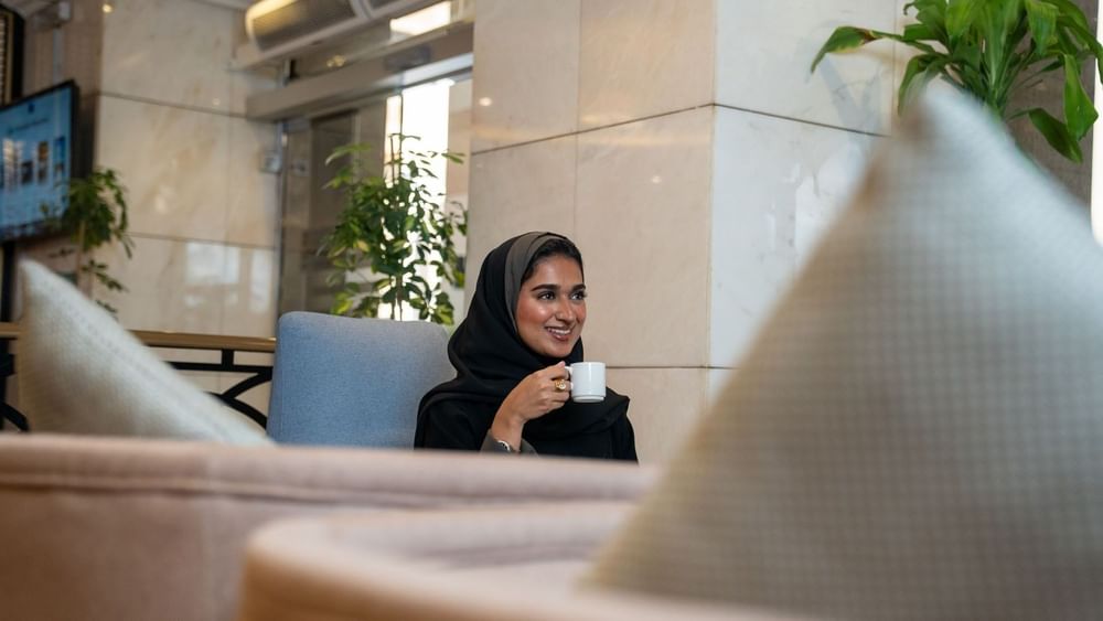 Woman wearing a black hijab smiling while holding a small coffee cup in Oasis Coffee Shop at Saja Warwick Madinah