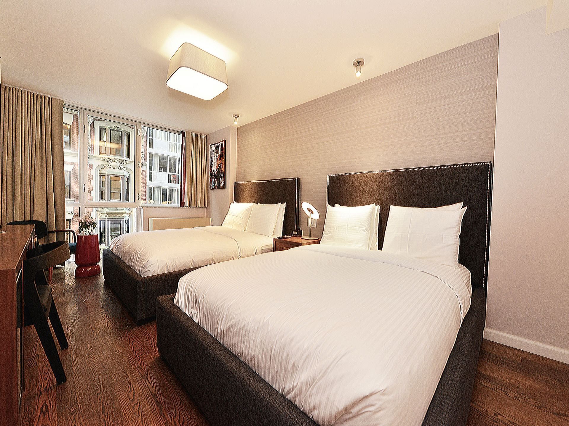 A hotel room with two beds, a desk, and a window showing a city view.