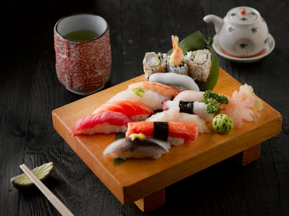 A traditional wooden sushi board with ten slices of colorful nigiri, a ball of wasabi, and a small pile of ginger, set on a dark table with a teapot, teacup, and a set of chopsticks.  