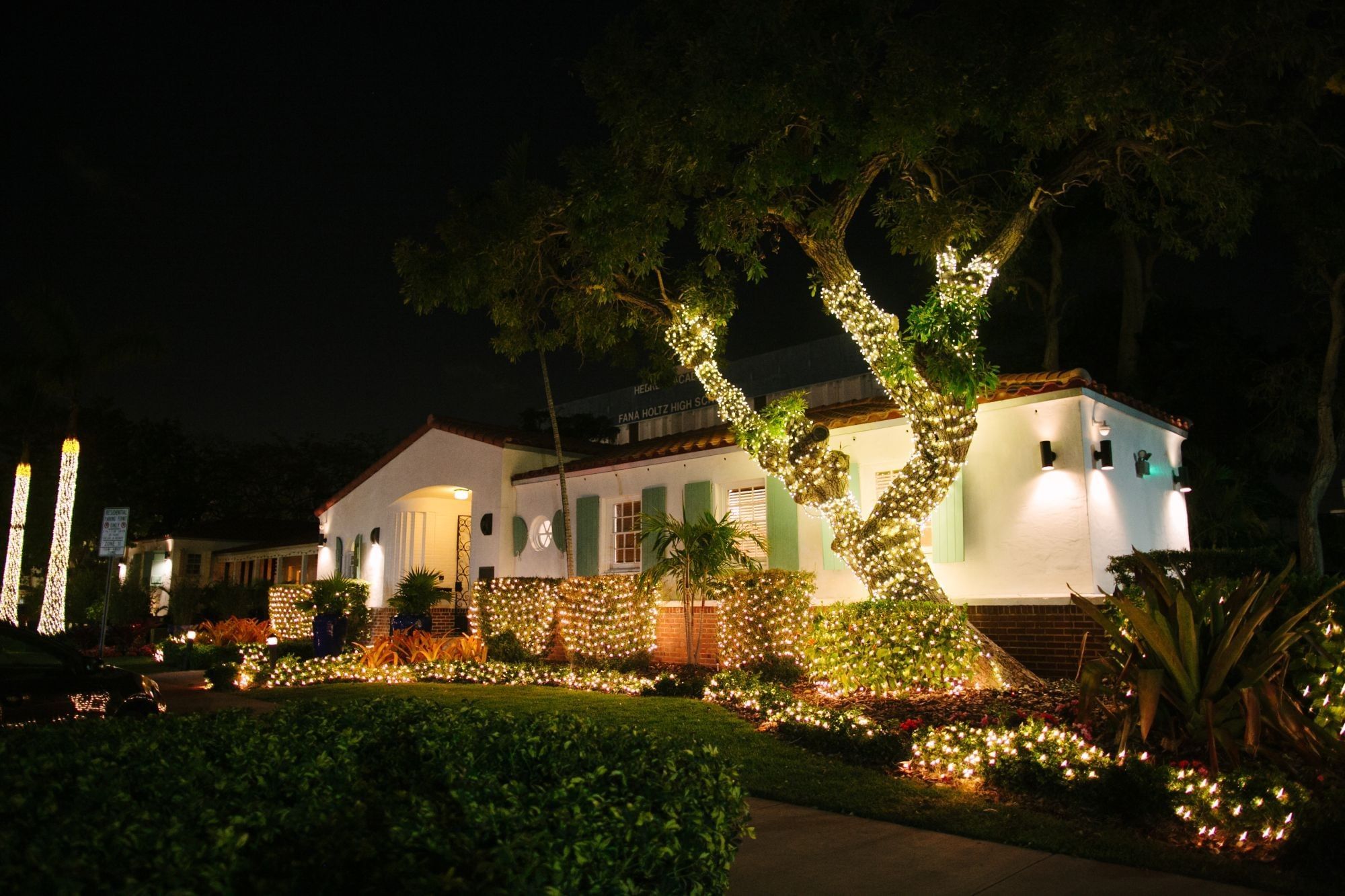 A house with white walls and green shutters is decorated with lights at night.