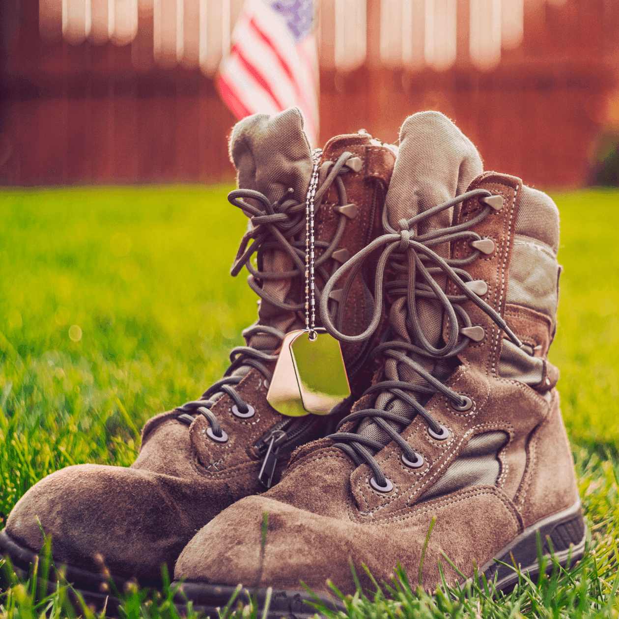 Pair of military boots with dog tags on grass, American flag in background.