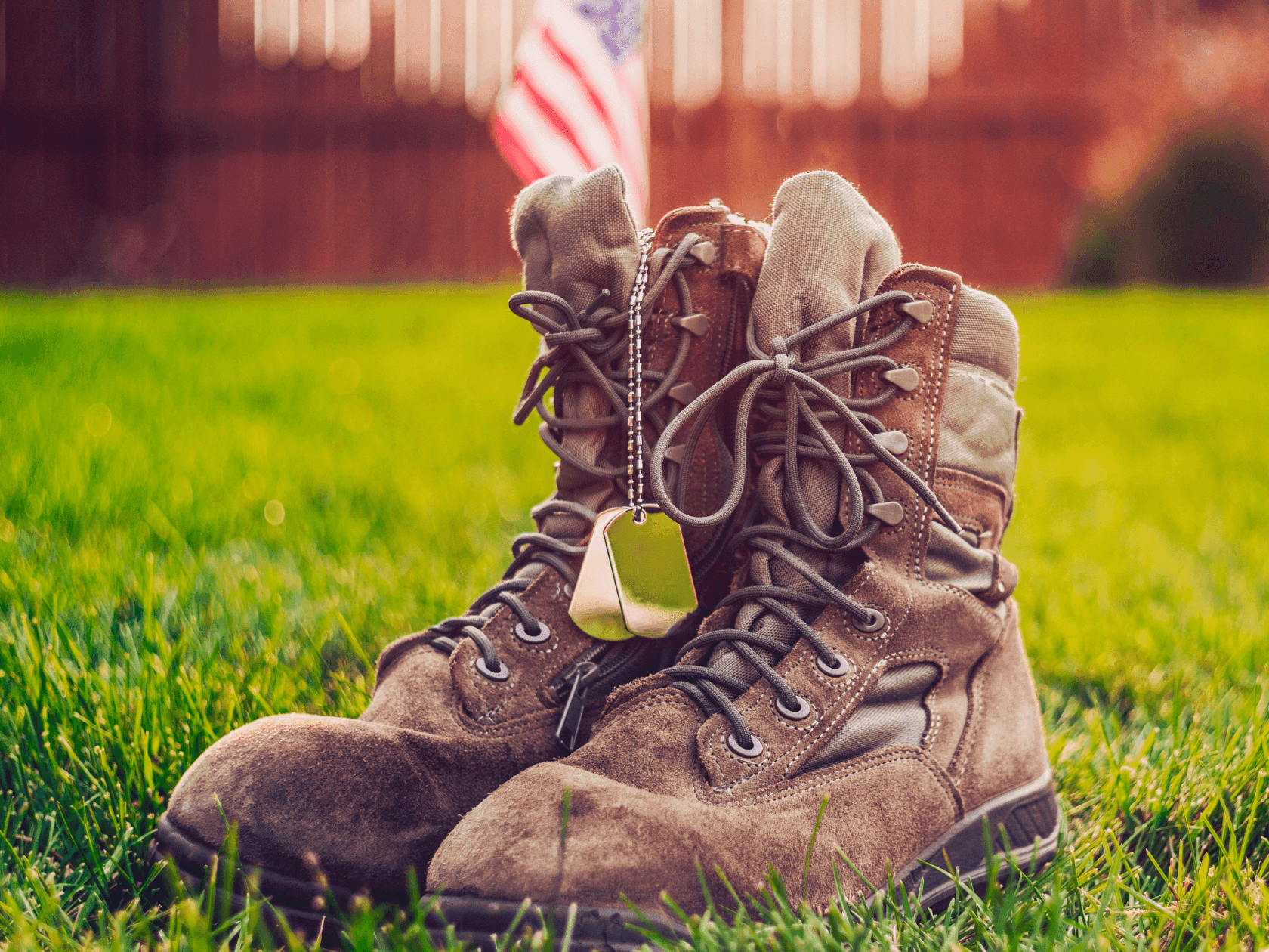 Pair of military boots on grass with dog tag, US flag in background.