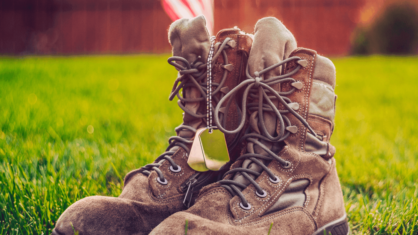 Pair of military boots with dog tags on grass, American flag in background.