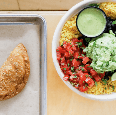 An empanada on a tray beside a bowl with fresh rice, tomatoes, and sauces from Black Bean Deli. 