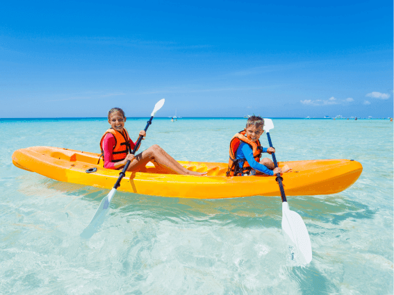Two smiling children in orange life vests paddle a bright yellow kayak in the ocean near The Plymouth Hotel South Beach