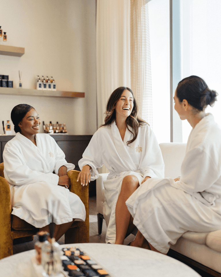 Three women in white spa robes laughing together, seated on gold and white chairs by a marble table at Hotel X Toronto