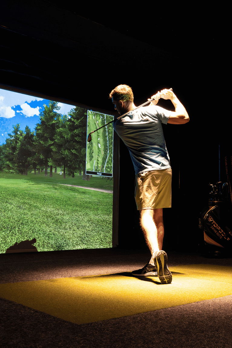 Golfer swinging a club on a simulator mat by a projected green course screen in the golf suite at Hotel X Toronto