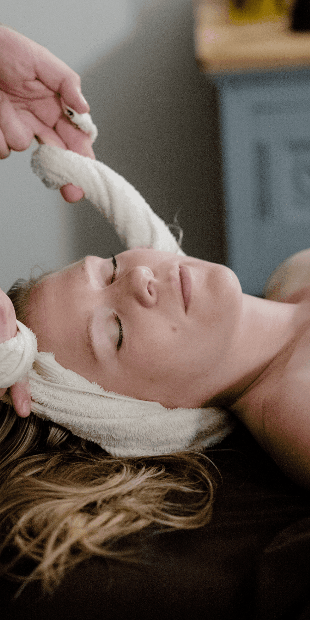 Esthetician holding a rolled warm towel above a guest\'s face resting on a spa treatment table at La Tourelle Inn & Spa
