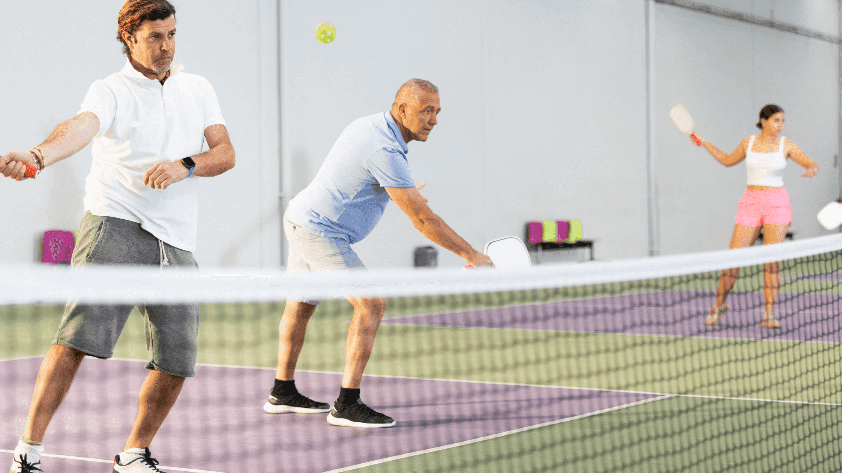 Three people play pickleball indoors, with two men on one side and a woman on the other.