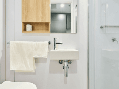 White bathroom with toilet, small sink, wooden cabinet, mirror, and glass shower enclosure.