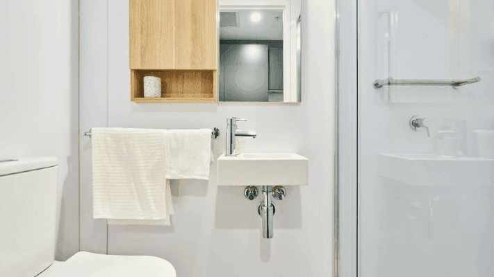 White bathroom with toilet, small sink, wooden cabinet, mirror, and glass shower enclosure.